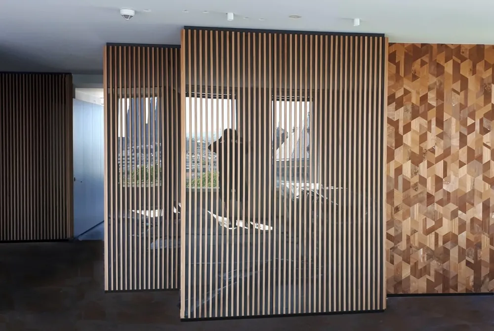 Raumteiler aus senkrechten Holzlamellen vor einer Fensterfront